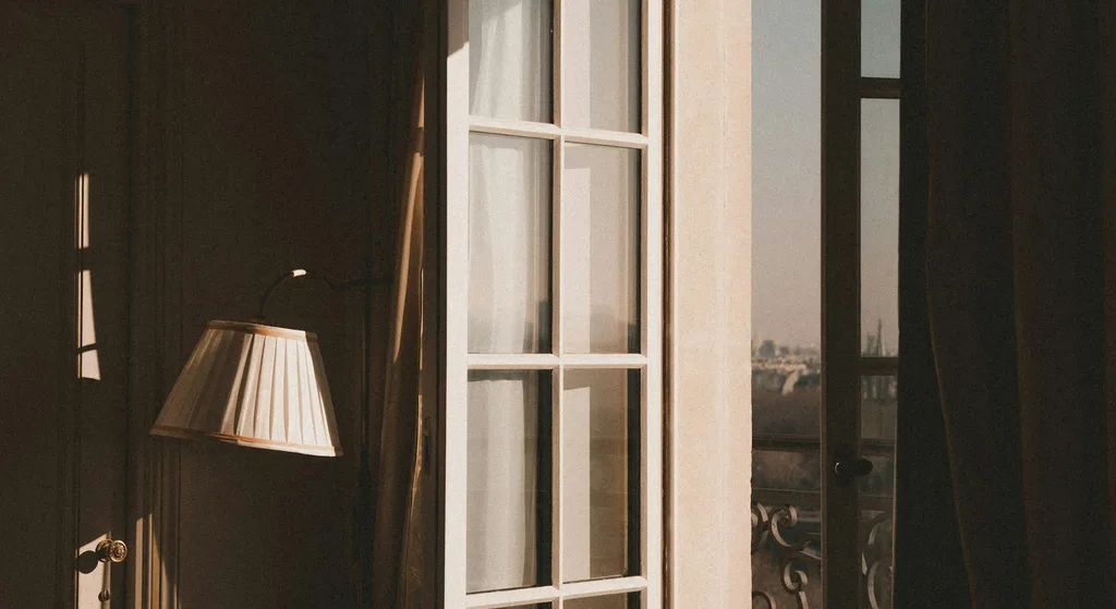 Warm evening light through an open hotel room window, with a lit floor lamp beside sheer curtains and a city skyline visible outside