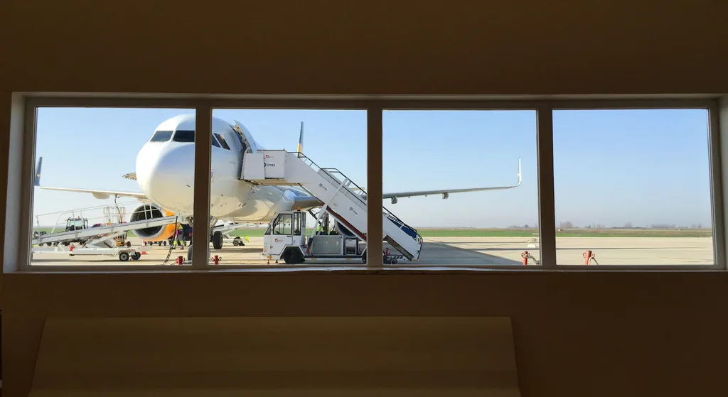 View through an airport terminal window of a passenger jet on the tarmac with a boarding staircase in place and ground crew working nearby